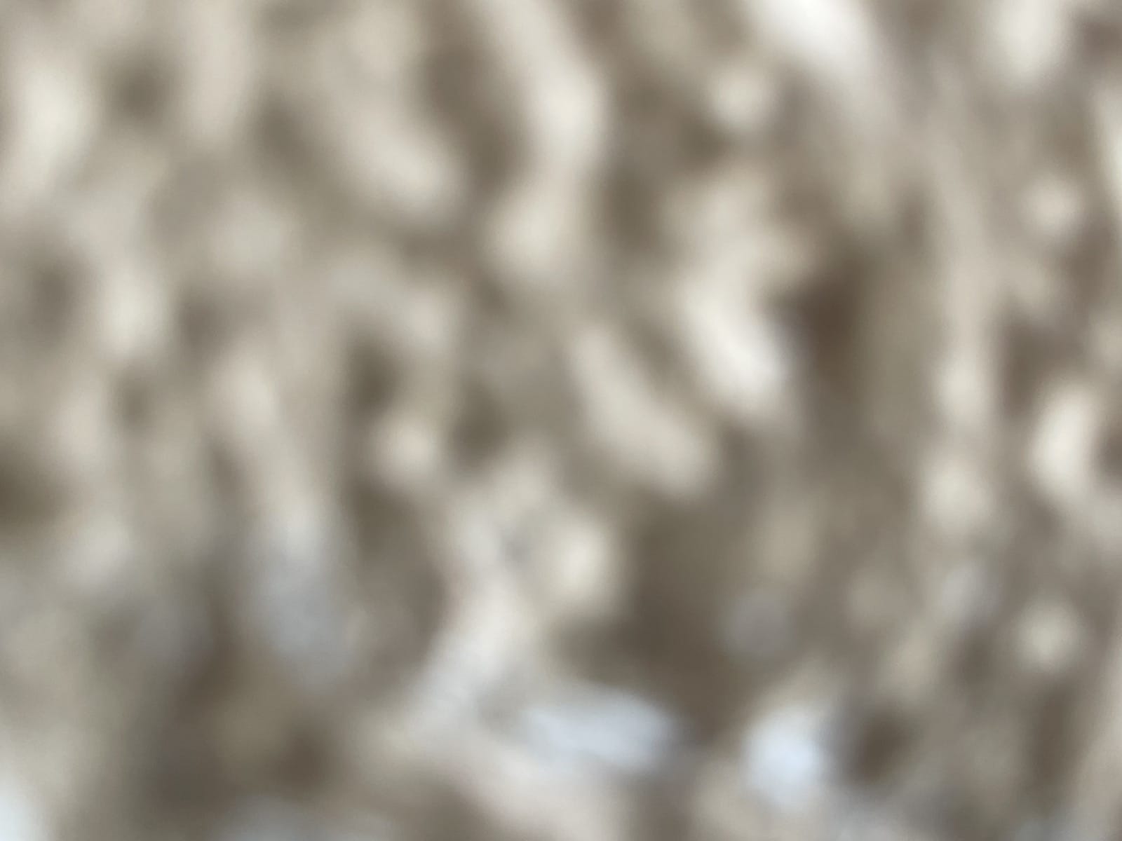 An out-of-focus, abstract close-up image featuring a textured surface in shades of beige, cream, and taupe. The bokeh effect creates soft, rounded shapes and a blurred, organic pattern reminiscent of carpet or grains of sand.